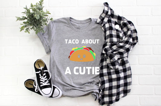 Grey t-shirt with white writing that says Taco About A Cutie. In between the writing is a smiling brown taco with brown, red, yellow and green coming out of it.