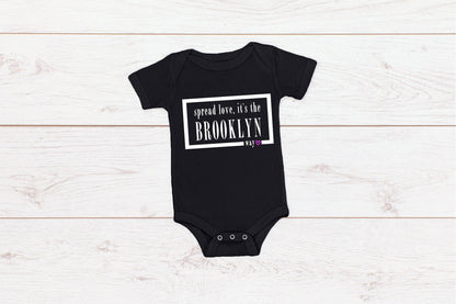 Black baby onesie with white writing that says spread love the Brooklyn way. Writing is surrounded by a split white box with the word way and a love heart to the bottom