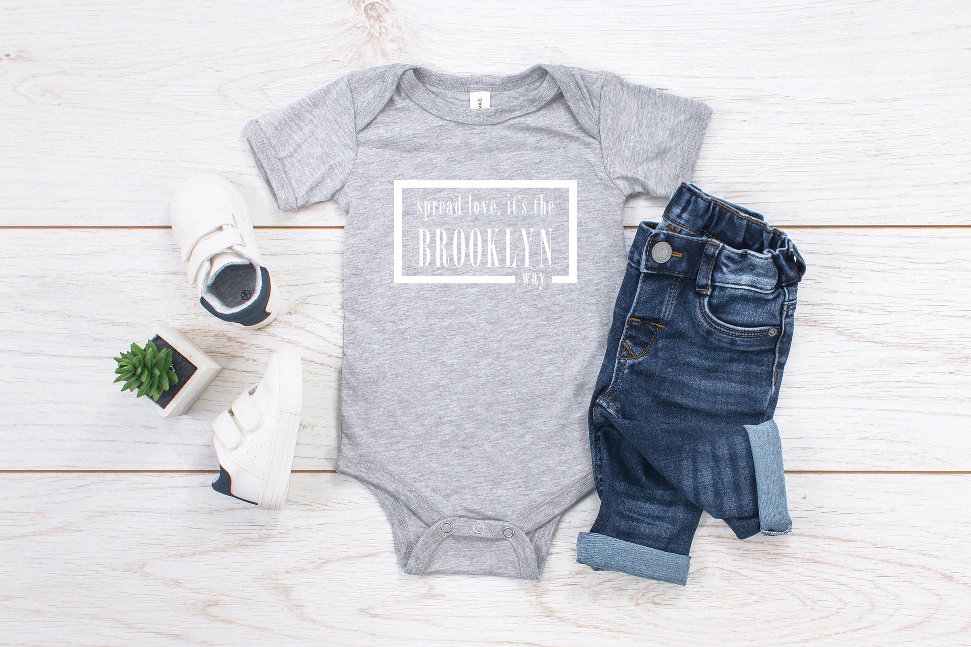 Grey baby romper that has white writing with spread love, it's the Brooklyn way. Surrounded by a white solid box.
